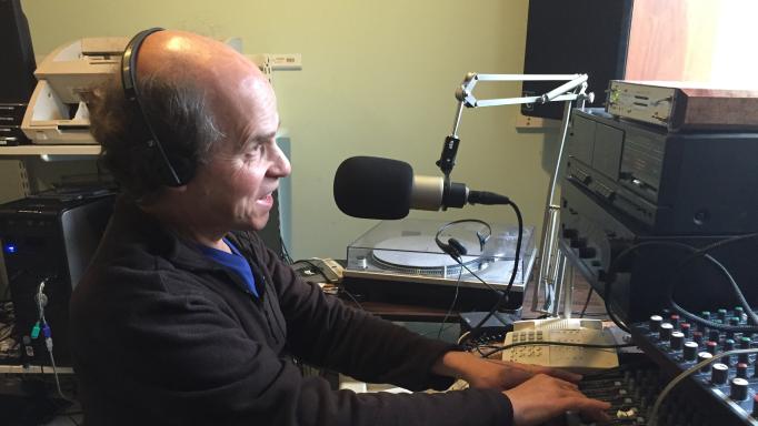 Photo of Glen Gordon in a recording studio.  He is wearing headphones, speaking into a microphone, and working with a mixer.